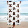 A person standing on a sandy beach holds up a large white blanket or towel featuring a repeating pattern of a teddybear bulldog. The ocean and blue sky are visible in the background.