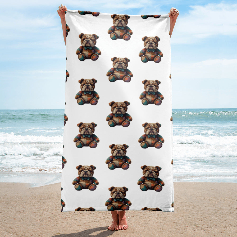 A person standing on a sandy beach holds up a large white blanket or towel featuring a repeating pattern of a teddybear bulldog. The ocean and blue sky are visible in the background.